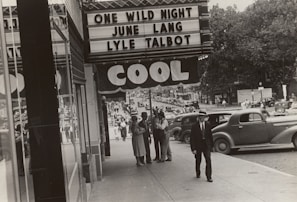 A black-and-white photograph depicts a bustling city street scene outside a theater. The marquee advertises the movie 'One Wild Night' featuring June Lang and Lyle Talbot. Several people, dressed in vintage clothing, walk along the sidewalk and converse in groups. Classic cars are parked along the street under the canopy bearing the word 'COOL'. Trees and various storefronts line the background, adding depth to the urban setting.