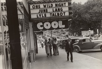 A black-and-white photograph depicts a bustling city street scene outside a theater. The marquee advertises the movie 'One Wild Night' featuring June Lang and Lyle Talbot. Several people, dressed in vintage clothing, walk along the sidewalk and converse in groups. Classic cars are parked along the street under the canopy bearing the word 'COOL'. Trees and various storefronts line the background, adding depth to the urban setting.