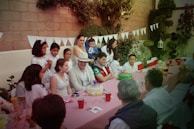 A group of people gathered around a long table set with a red-checkered tablecloth, celebrating in an outdoor garden setting. Various adults and children are present, some standing and others seated, with decorated cakes and drinks on the table. The background features greenery and decorative white bunting hanging along a brick wall, contributing to a festive atmosphere.