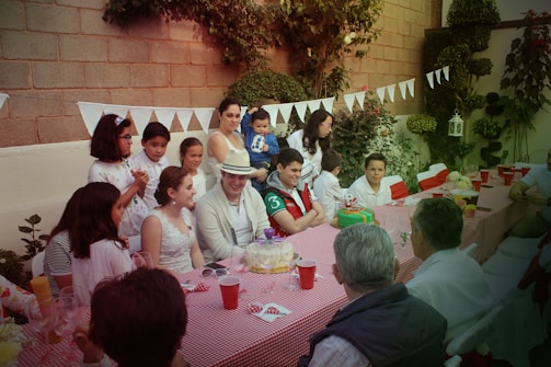 A group of people gathered around a long table set with a red-checkered tablecloth, celebrating in an outdoor garden setting. Various adults and children are present, some standing and others seated, with decorated cakes and drinks on the table. The background features greenery and decorative white bunting hanging along a brick wall, contributing to a festive atmosphere.