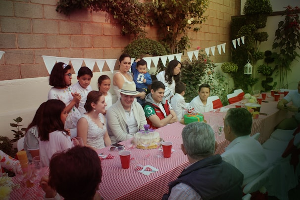 A group of people gathered around a long table set with a red-checkered tablecloth, celebrating in an outdoor garden setting. Various adults and children are present, some standing and others seated, with decorated cakes and drinks on the table. The background features greenery and decorative white bunting hanging along a brick wall, contributing to a festive atmosphere.