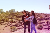 Two people are standing in an archaeological site with stone terraces and scattered palm trees. The individuals appear to be enjoying the scenery, with an expansive view of the site behind them. The landscape is sunlit, providing a warm and inviting atmosphere.