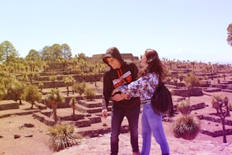 Two people are standing in an archaeological site with stone terraces and scattered palm trees. The individuals appear to be enjoying the scenery, with an expansive view of the site behind them. The landscape is sunlit, providing a warm and inviting atmosphere.