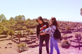 Two people are standing in an archaeological site with stone terraces and scattered palm trees. The individuals appear to be enjoying the scenery, with an expansive view of the site behind them. The landscape is sunlit, providing a warm and inviting atmosphere.