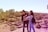 Two people are standing in an archaeological site with stone terraces and scattered palm trees. The individuals appear to be enjoying the scenery, with an expansive view of the site behind them. The landscape is sunlit, providing a warm and inviting atmosphere.