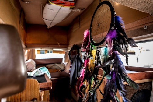 The interior of a van features a colorful dreamcatcher with feathers in shades of pink, purple, blue, yellow, and black. A surfboard is mounted on the ceiling while assorted fabrics and a blanket are seen on the bed in the background.