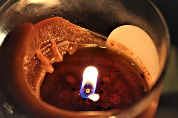 A close-up view of a lit candle with partially melted wax. The flame is small, and the wick is visible, surrounded by a smooth, glossy surface of solidified wax. The edges of the wax appear uneven and textured.