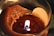 A close-up view of a lit candle with partially melted wax. The flame is small, and the wick is visible, surrounded by a smooth, glossy surface of solidified wax. The edges of the wax appear uneven and textured.