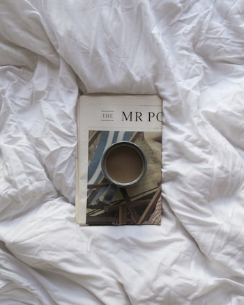 A magazine lies on a rumpled white bedspread with a cup of coffee placed on top of it. The magazine cover has a partial image of beach chairs and a printed title. The setting suggests a relaxed morning atmosphere.