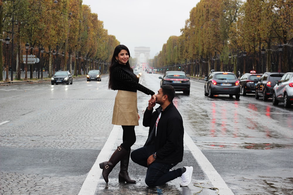 man wearing black jacket kneeling in front of woman kissing hand on road
