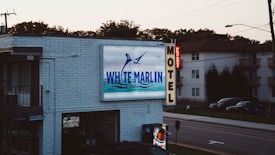 A motel sign displays the name 'White Marlin Inn' with an image of a marlin fish above blue ocean waves. The word 'Motel' is vertically displayed on a bright sign, which includes a smaller neon 'Vacancy' sign. In the background, there are residential buildings and parked cars along a street, with trees lining the horizon.