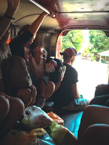 Passengers are seated inside a vehicle, likely a jeepney, with a child resting on an adult's lap. Bags are placed on the floor and sunlight is streaming in from the back, illuminating the greenery and street outside.