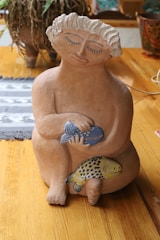 A clay sculpture of a person with closed eyes and slightly smiling lips, holding two colorful fish against a wooden background. The sculpture features intricate detailing with curly hair and a serene expression.