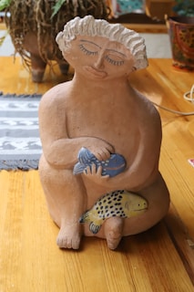 A clay sculpture of a person with closed eyes and slightly smiling lips, holding two colorful fish against a wooden background. The sculpture features intricate detailing with curly hair and a serene expression.