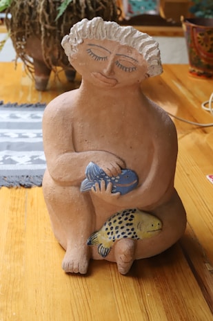 A clay sculpture of a person with closed eyes and slightly smiling lips, holding two colorful fish against a wooden background. The sculpture features intricate detailing with curly hair and a serene expression.
