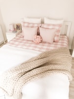 A neatly made bed featuring a pastel pink and white checkered duvet with multiple pillows arranged against a white headboard. A chunky beige knit blanket is draped over the bed. On the nightstand, there are small decorative items including books and a small framed photo.