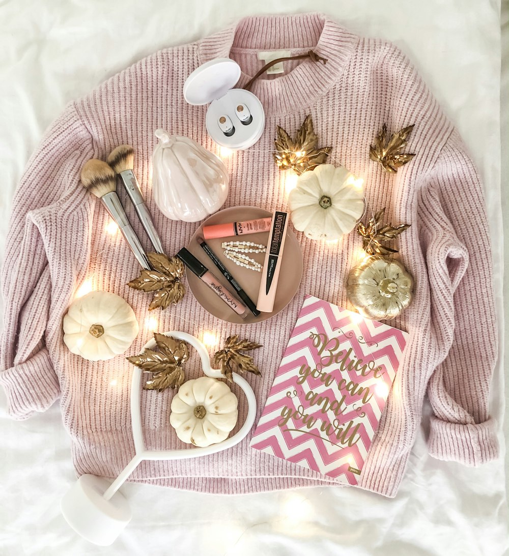 white pumpkin, lipsticks, gold leaves, makeup brush, and pink and white paper bag on pink crew-neck sweater