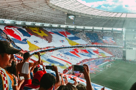 A vibrant soccer stadium filled with enthusiastic fans. Giant banners and flags with various colors and symbols cover a large section of the stands. The crowd is dense and actively engaged, some holding up phones to capture the moment.