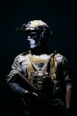 A person dressed in tactical military gear stands against a dark background. The attire includes a helmet with mounted equipment, a skull-patterned face mask, and a tactical vest with various attachments. The lighting creates a dramatic contrast, highlighting the figure's gear and muscular build.