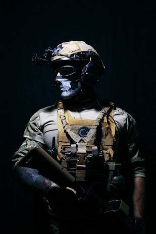 A person dressed in tactical military gear stands against a dark background. The attire includes a helmet with mounted equipment, a skull-patterned face mask, and a tactical vest with various attachments. The lighting creates a dramatic contrast, highlighting the figure's gear and muscular build.