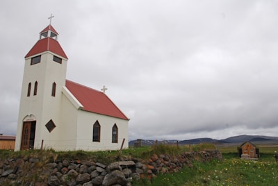 A small white church with a red roof stands prominently in a rural landscape. The church features a tall steeple with a cross on top, and several arched windows. A low stone wall runs along a grassy area dotted with wildflowers. In the background, there are rolling hills and cloudy skies, creating a tranquil, isolated atmosphere.