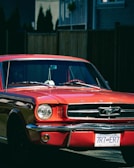 A red vintage car with a reflective chrome grille is parked in front of a wooden fence in a residential area. The license plate indicates a location in British Columbia, and a small ornament hangs from the rearview mirror inside the car.