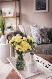 A stylish, cozy living room features a bouquet of yellow flowers in a glass jar on a decorative white tray. The background includes a comfortable sofa with a patterned pillow, a potted plant, and neatly arranged shelves with framed photos.