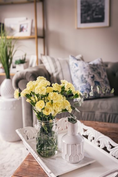 A stylish, cozy living room features a bouquet of yellow flowers in a glass jar on a decorative white tray. The background includes a comfortable sofa with a patterned pillow, a potted plant, and neatly arranged shelves with framed photos.