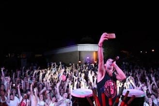 A lively concert scene with a large crowd of enthusiastic people raising their hands, illuminated by colorful lights. A performer is in the foreground taking a selfie with a smartphone while holding a microphone, creating an energetic and engaging atmosphere.