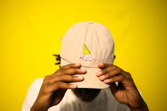 A person wearing a beige baseball cap with an ice cream cone graphic on the front. The bright yellow background contrasts with the cap. The person's hands are adjusting the cap, which partially obscures their face.