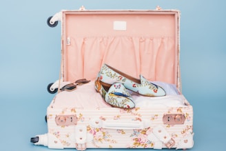 A floral patterned suitcase is open, revealing a pair of stylish loafers with a floral and bird design placed on top of neatly packed clothes. Nearby, a pair of sunglasses rests casually. The interior of the suitcase is lined with a soft pink fabric.