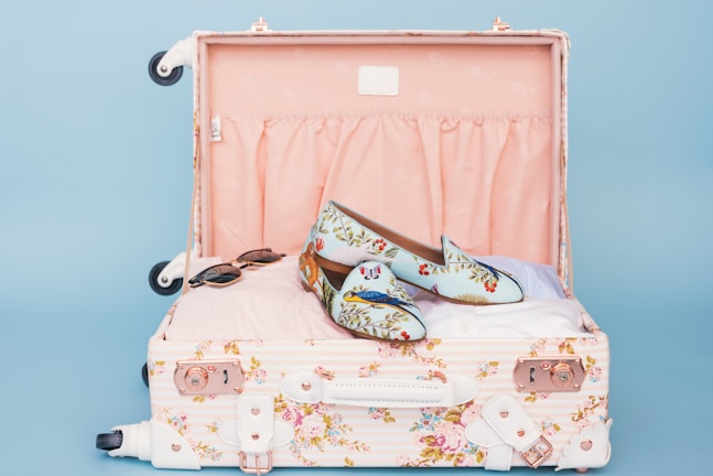 A floral patterned suitcase is open, revealing a pair of stylish loafers with a floral and bird design placed on top of neatly packed clothes. Nearby, a pair of sunglasses rests casually. The interior of the suitcase is lined with a soft pink fabric.