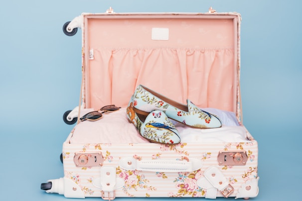 A floral patterned suitcase is open, revealing a pair of stylish loafers with a floral and bird design placed on top of neatly packed clothes. Nearby, a pair of sunglasses rests casually. The interior of the suitcase is lined with a soft pink fabric.