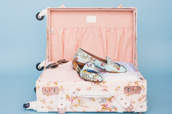 A floral patterned suitcase is open, revealing a pair of stylish loafers with a floral and bird design placed on top of neatly packed clothes. Nearby, a pair of sunglasses rests casually. The interior of the suitcase is lined with a soft pink fabric.