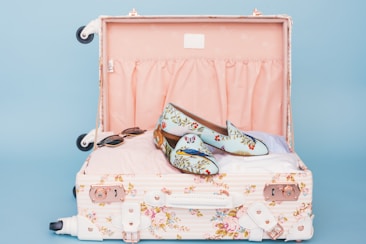 A floral patterned suitcase is open, revealing a pair of stylish loafers with a floral and bird design placed on top of neatly packed clothes. Nearby, a pair of sunglasses rests casually. The interior of the suitcase is lined with a soft pink fabric.