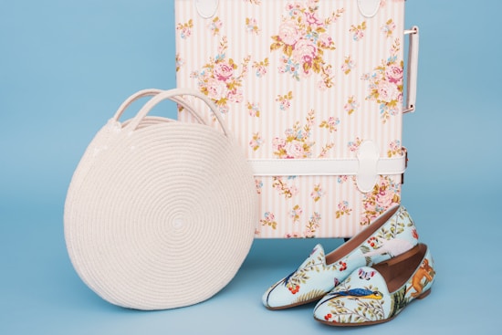 A pastel floral suitcase sits against a light blue background, accompanied by a round white woven bag and a pair of slip-on shoes decorated with colorful bird and floral patterns.