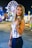 A young woman with long hair stands in the foreground of a nighttime carnival setting. Behind her, a brightly lit Ferris wheel and colorful carnival booths create a vibrant backdrop. She is wearing a blue and white tie-dye top and jeans, accessorized with bracelets. The overall scene has a lively and festive atmosphere.