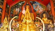 A richly decorated shrine houses several golden Buddha statues of varying sizes, surrounded by intricate, brightly colored murals depicting scenes of traditional life and spiritual imagery. Floral arrangements and symbolic objects accentuate the setting, creating a vibrant and serene atmosphere.