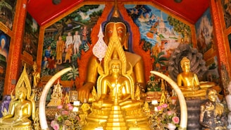 A richly decorated shrine houses several golden Buddha statues of varying sizes, surrounded by intricate, brightly colored murals depicting scenes of traditional life and spiritual imagery. Floral arrangements and symbolic objects accentuate the setting, creating a vibrant and serene atmosphere.