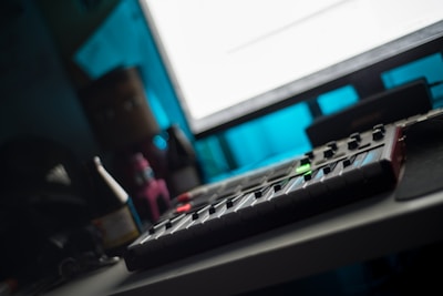 A dimly lit room with a music production setup including a MIDI controller on a desk, surrounded by bottles and other objects. A large monitor casts a bright glow over the scene while providing a contrast to the otherwise dark environment.