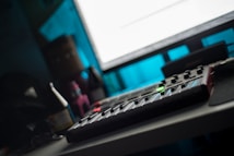 A dimly lit room with a music production setup including a MIDI controller on a desk, surrounded by bottles and other objects. A large monitor casts a bright glow over the scene while providing a contrast to the otherwise dark environment.