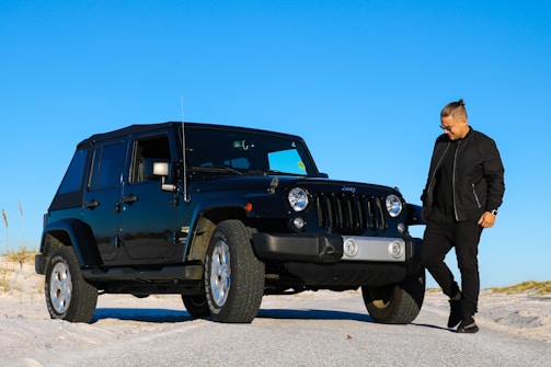 A person wearing a black jacket and sunglasses stands beside a black Jeep parked on a sandy terrain with a clear blue sky above.