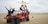 A group of eight people are posing on and around an orange dune buggy in a desert setting. Some are sitting and others are standing, with expressions of joy and excitement as they make peace signs and gestures. The landscape features expansive sand dunes under a cloudy sky.