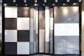 An arrangement of large vertical panels featuring various textures and colors, including black and white checkered patterns, solid gray, solid white, and a natural wood pattern. The panels are backlit by spotlights mounted on the ceiling, creating shadows and highlights on their surfaces.