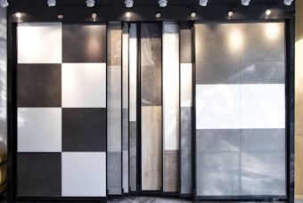 An arrangement of large vertical panels featuring various textures and colors, including black and white checkered patterns, solid gray, solid white, and a natural wood pattern. The panels are backlit by spotlights mounted on the ceiling, creating shadows and highlights on their surfaces.