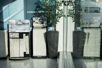 Several tile samples are displayed on stands with various designs visible, situated against a blue wall with text in the background. Large potted plants are placed beside the displays, and a glass reflection adds depth to the scene.