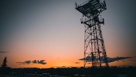 A silhouetted communication tower stands prominently against a vibrant sunset backdrop, with hues of orange and yellow filling the sky. Dark clouds are scattered across the horizon, and a few trees are visible in the distance.