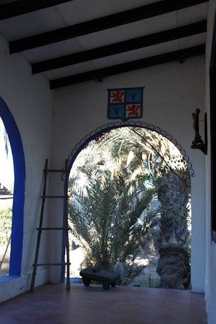 A rustic outdoor veranda with a view of lush palm trees. The area has a simple ladder against a white wall with blue accents, and a decorative crest above the archway. There is a traditional bench or small table in the foreground.