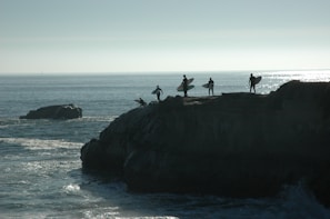 Four surfers holding their surfboards stand on a cliff overlooking the ocean. The silhouette of their figures is sharply contrasted against the bright, shimmering water in the background. The scene conveys a sense of anticipation and adventure as they prepare to surf.