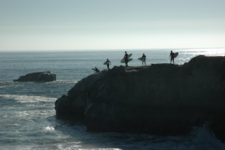 Four surfers holding their surfboards stand on a cliff overlooking the ocean. The silhouette of their figures is sharply contrasted against the bright, shimmering water in the background. The scene conveys a sense of anticipation and adventure as they prepare to surf.
